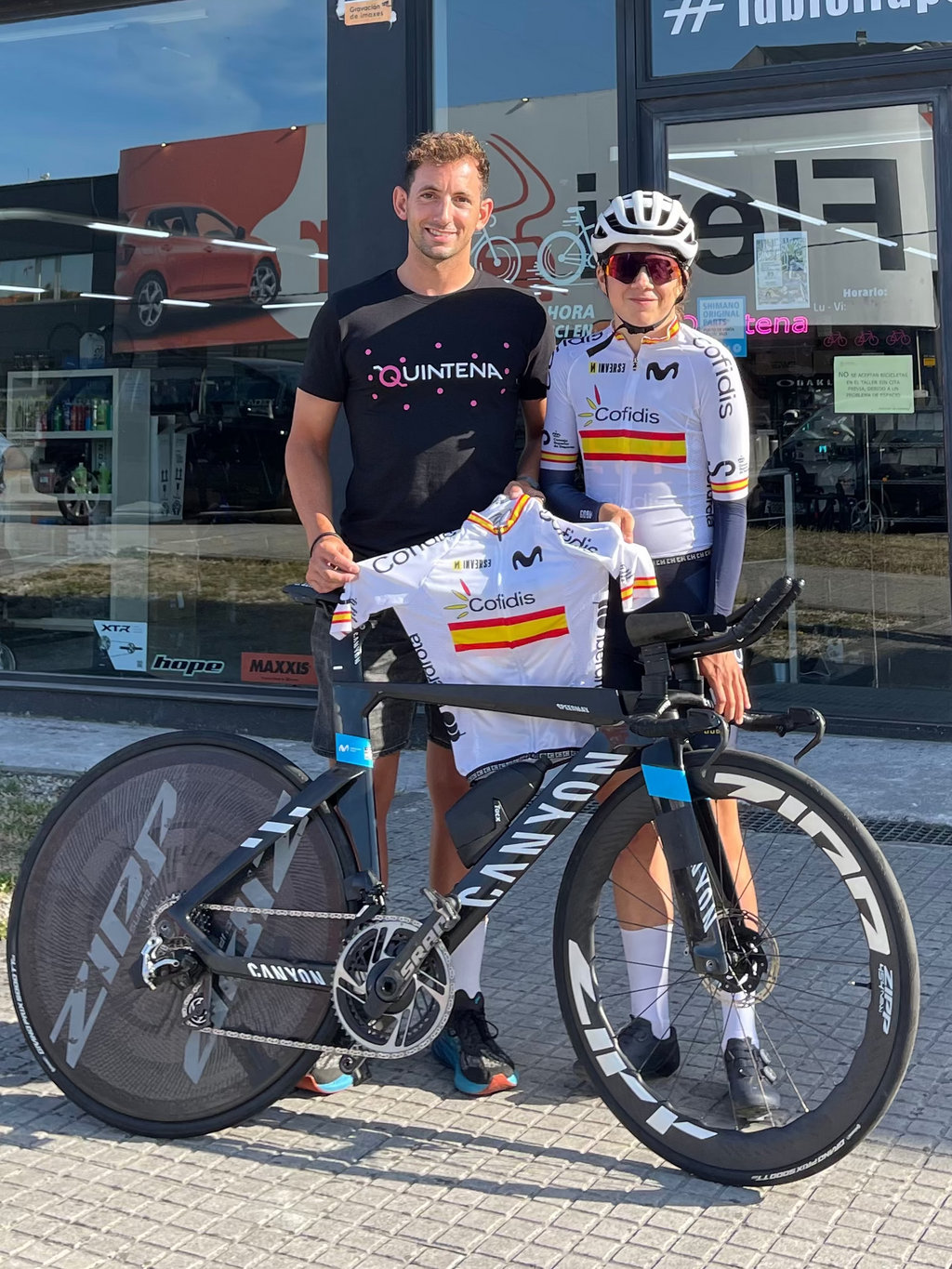 BikeFitter and Cyclist from the movistar team posing with a black TT bicycle in front of the Bike Fit Center storefront under a blue sky with the jersey of national champion.