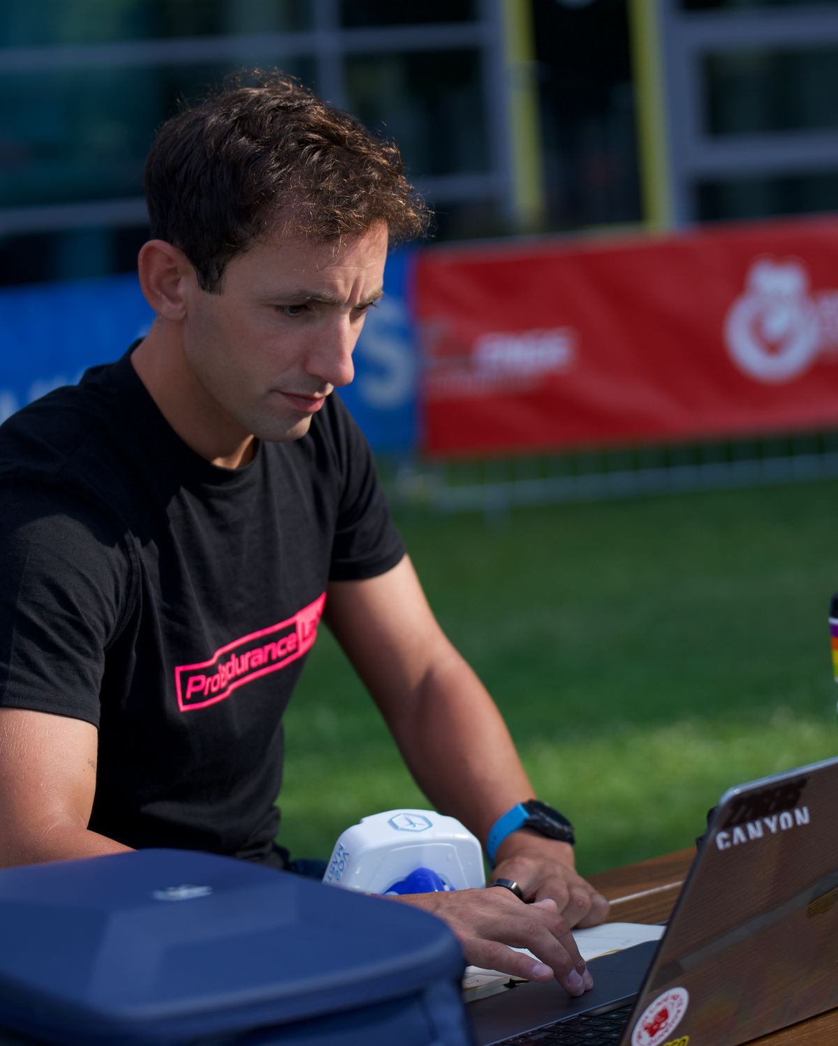 A man wearing a black ProRide t-shirt sits at a table outdoors using a laptop. A black Canyon bike bag and a black Master-R case are on the table next to him, with a grassy field and event banners in the background.